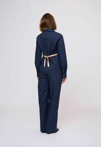 Tengerészkék, hosszú ujjú ing, húzott hátoldallal és bézs szalaggal megkötve deréknál, hozzá illő, bő szárú nadrággal. Simább textúrájú anyag.