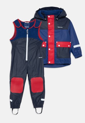 Marineblaue Regenjacke mit roten und blauen Akzenten, ausgestattet mit einem Front-Reißverschluss und Druckknöpfen. Passende marineblaue Latzhose mit roten Kniewohlfühlstellen.