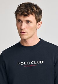 Marinblå sweatshirt med rund halsringning. Har "POLO CLUB" och "ETABLERAD 2012" tryckt i vit och röd text över bröstet.