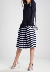 Haut à manches longues bleu marine avec texture plissée, associé à une jupe trapèze rayée bleu marine et blanche. Sandales à talons argentées et petit sac argenté.