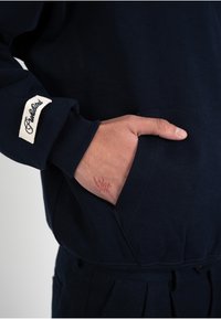 Main partiellement dans la poche avant d'un sweat à capuche bleu marine avec un patch blanc sur la manche portant l'inscription « Established » en écriture cursive.