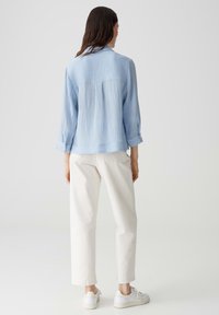 Camicia a maniche lunghe azzurro chiaro, con texture, colletto e polsini con bottoni, abbinata a pantaloni bianchi a gamba dritta e sneakers bianche.