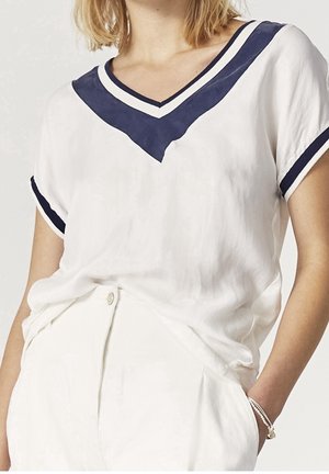 Femme portant un t-shirt blanc à col en V avec un liseré bleu marine sur le col et les manches, associé à un pantalon blanc et un bracelet argenté au poignet droit.