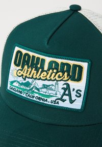 Grønn snapback-cap med et rektangulært merkelapp som har "Oakland Athletics", en fjellgrafikk og teksturer i stoffet og sømmen.