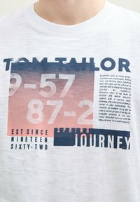 Valkoinen puuvillateepaita, jossa on graafinen printti sinisellä ja vaaleanpunaisella tekstillä, mukaan lukien "TOM TAILOR" ja numeroita sekä "SPRING JOURNEY" -yksityiskohtia.