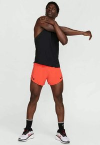 Schwarzes ärmelloses Oberteil, orange Shorts mit einem Logo und weiße Sportschuhe. Die Person streckt einen Arm über ihren Körper.