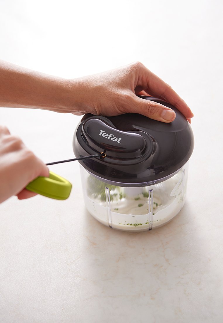 Tirant sur la corde d'un hachoir manuel Tefal noir contenant de la crème blanche avec des herbes vertes sur un plan de travail clair.