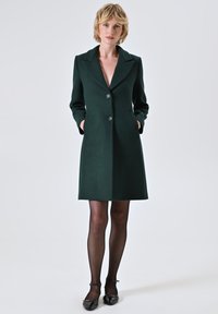 Cappotto di lana verde scuro con collo a revers, due bottoni e tasche laterali. Vestibilità sartoriale, lunghezza al ginocchio, abbinato a collant neri e scarpe.