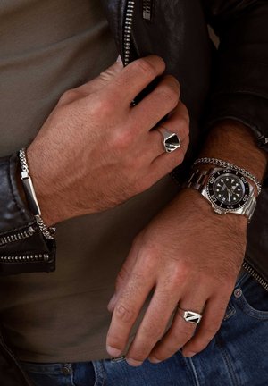 Silver chain bracelet, silver ring with geometric design, and a stainless steel watch with black dial, worn on a hand against a leather jacket.