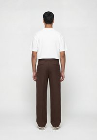 Άνδρας όρθιος με πλάτη, φοράει λευκό πουκάμισο με κοντά μανίκια, καφέ φαρδιές παντελόνες και λευκά αθλητικά παπούτσια, σε απλό ανοιχτό φόντο.