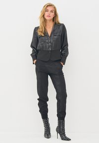 Juoda satino palaidinė su trumpu dizainu, ilgomis rankovėmis ir sagų detalėmis, derinama su juodomis siaurėjančiomis kelnėmis su tekstūruotais akcentais ir auliniais batais su kniedėmis.