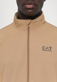 Šviesiai rudas džemperis su aukštu apykaklės, užtrauktuku ir EA7 Emporio Armani logotipu. Lygus paviršius, lengvas audinys.