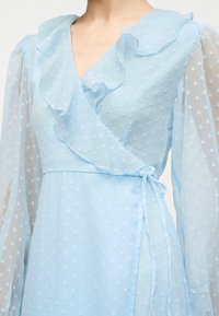 Φόρεμα με ελαφρώς μπλε απόχρωση, με διαφανή μακριά μανίκια, υφή με πουά, V-λαιμόκοψη με βολάν και κλείσιμο με κορδέλα στο πλάι.