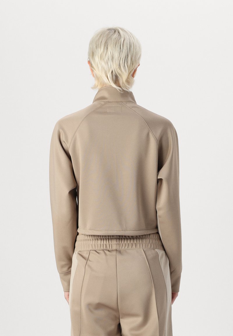 Giacca atletica tagliata beige con colletto alto, maniche lunghe e una texture liscia. Presenta dettagli con cintura elastica e un design minimalista.