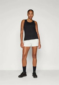 Crna majica bez rukava uparena s bijelim kratkim hlačama. Model nosi crne tenisice i tamnoplave čarape. Glatka tkanina, uski kroj.