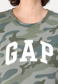 Zelené maskáčové tričko vyrobené z bavlny, s kulatým výstřihem a výrazným bílým nápisem "GAP" na prsou. Hladký materiál s jemnými vzory.