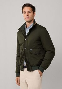 Uomo che indossa una giacca verde scuro con bottoni sopra una camicia a righe e pantaloni beige, in piedi con una mano in tasca su uno sfondo semplice.