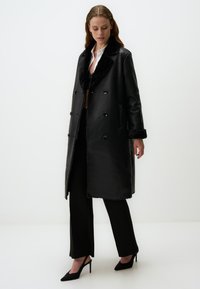 Manteau noir en cuir avec col en fausse fourrure, design à double boutonnage et poches latérales. Modèle assorti avec un pantalon noir et des talons.