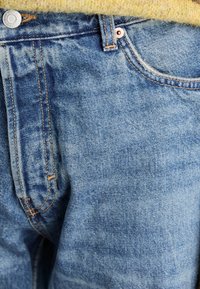 Jeans de mezclilla en lavado azul claro con costuras visibles, herrajes de cobre y un ligero patrón de desgaste. Presentan un cierre de botón y cremallera.