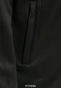 Prim-plan al unui articol de îmbrăcăminte din țesătură neagră, cu un buzunar vertical cu clapetă și cuvântul „BECOOL” imprimat aproape de bază.