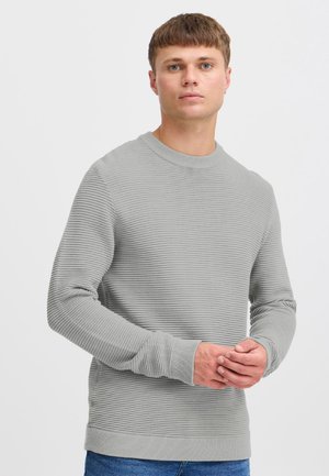 Giovane uomo con capelli castani corti che indossa un maglione grigio chiaro a costine e jeans blu, con le mani giunte davanti a uno sfondo bianco.