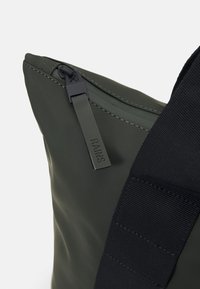 Mørkegrønn vanntett pose med glidelås, med en svart stropp og merkeetikett. Jevn tekstur og minimalistisk design.