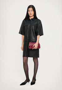 Γυναίκα που φοράει ένα μαύρο δερμάτινο φόρεμα, διάφανες μαύρες καλσόν, μαύρες γατούλες ψηλοτάκουνες, κρατώντας μία γυαλιστερή κόκκινη τσάντα χειρός.