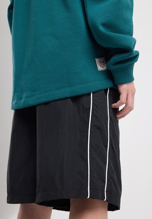 Personne portant un t-shirt à manches longues turquoise avec un écusson USA et un short noir avec un liseré blanc vertical, debout devant un fond uni.