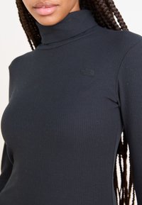 Camisola preta de gola alta, com manga longa e um design ajustado. Apresenta um pequeno logótipo em relevo no peito. Textura suave.