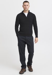 Zwarte gebreide trui met een ritskraag, voorzien van een gestructureerd patroon. Gepaard met donkere cargo-broeken en bruine schoenen met een patroon.