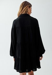 Abito nero caratterizzato da un design a strati, maniche lunghe e voluminose e un colletto. Il tessuto ha un aspetto testurizzato e stropicciato.