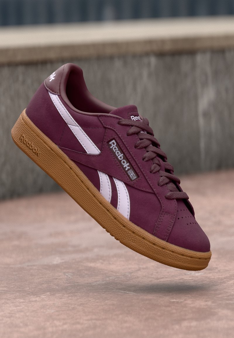 Sapatilha bordeaux com detalhes brancos, parte superior em camurça texturizada e sola de borracha natural. Apresenta atacadores e a marca Reebok na lateral.