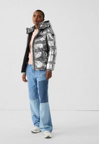 Zilveren pufferjack met capuchon, voorzien van doorgestikte gedeelten. Gecombineerd met blauwe patchwork spijkerbroek en lichte sneakers. Soepele textuur en glanzende afwerking.