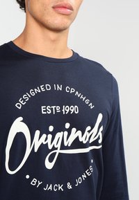 Navyfarbener Langarm-T-Shirt aus Baumwolle mit einem großen weißen Druck, der "Originals" lautet, sowie zusätzlichem Text darüber und darunter.