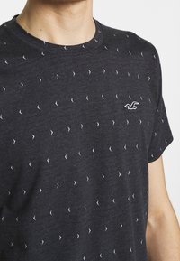 Tmavě šedé tričko s kulatým výstřihem, zdobené opakujícím se vzorem malých bílých šipek a bílým logem značky. Měkká, texturovaná tkanina.