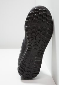 Musta tennarin pohja, jossa on teksturoitua kumia ja pitoelimiä, hieman kohotettu kanta ja sileät reunat.