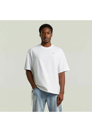 Uomo con maglietta bianca oversize e jeans azzurri, in piedi davanti a uno sfondo grigio chiaro uniforme, con una mano in tasca.