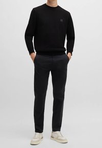 Sweat-shirt noir à col rond, avec un petit patch logo en relief, associé à un pantalon foncé et des baskets de couleur claire.