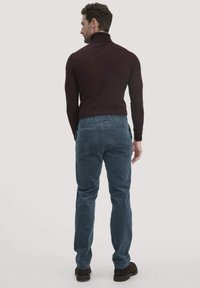 Burgunderfarbener Rollkragenpullover, taillierte Passform. Petrolblaue Cordhosen mit Gesäßtaschen. Modell wird von hinten gezeigt, trägt dunkle Schuhe.