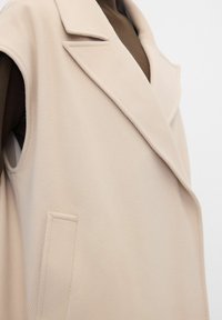 Beiger ärmelloser Mantel mit weitem Revers, getragen über einem braunen Langarmshirt, zeigt Textur und maßgeschneiderte Taschen-Details.