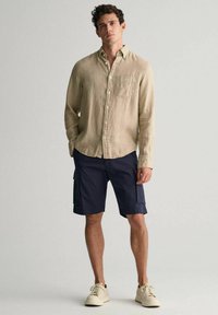 Beige Leinen-Hemd mit Knöpfen und einer Brusttasche, kombiniert mit navy farbenen Cargo-Shorts mit seitlichen Taschen und hellen Sneakers.