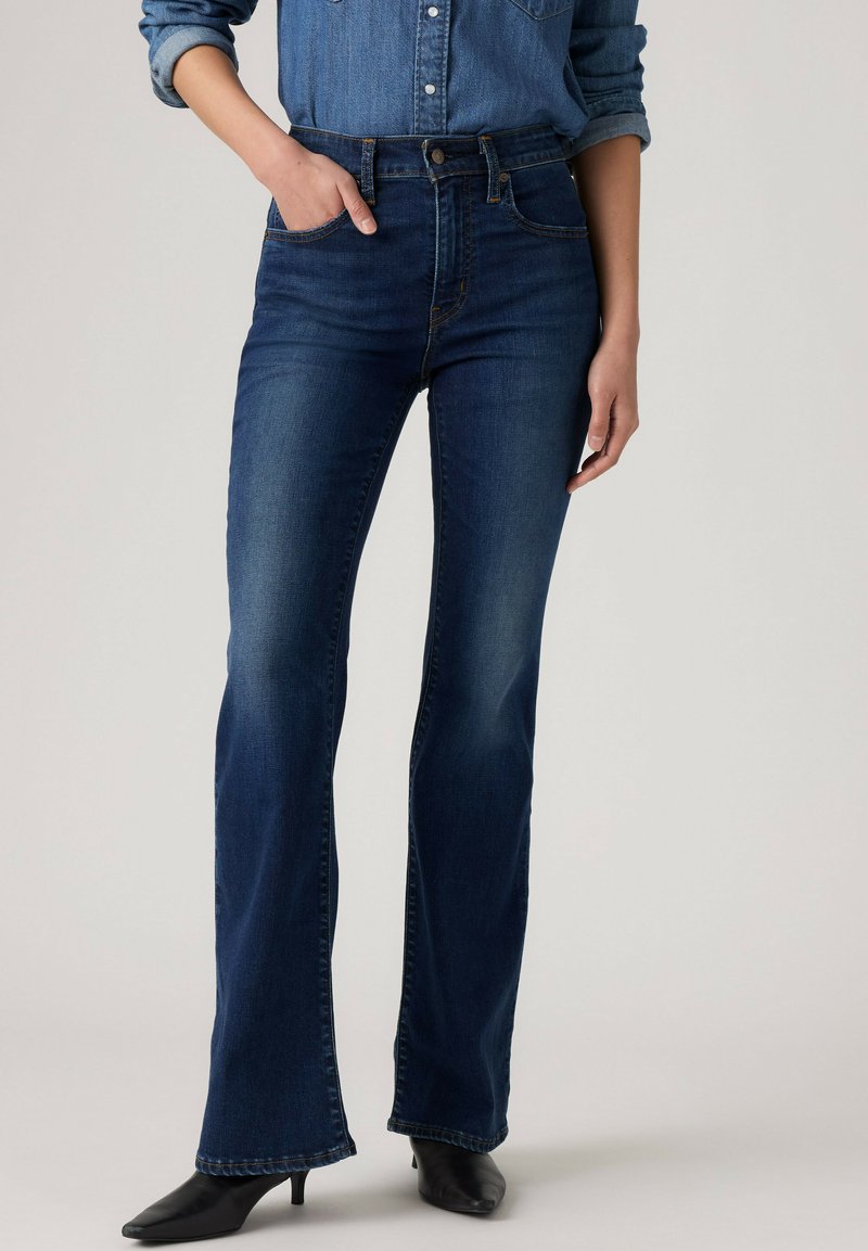Personne portant un jean bootcut bleu foncé, une main dans la poche, associée à des talons noirs à bout pointu et une chemise en denim manches longues retroussées.