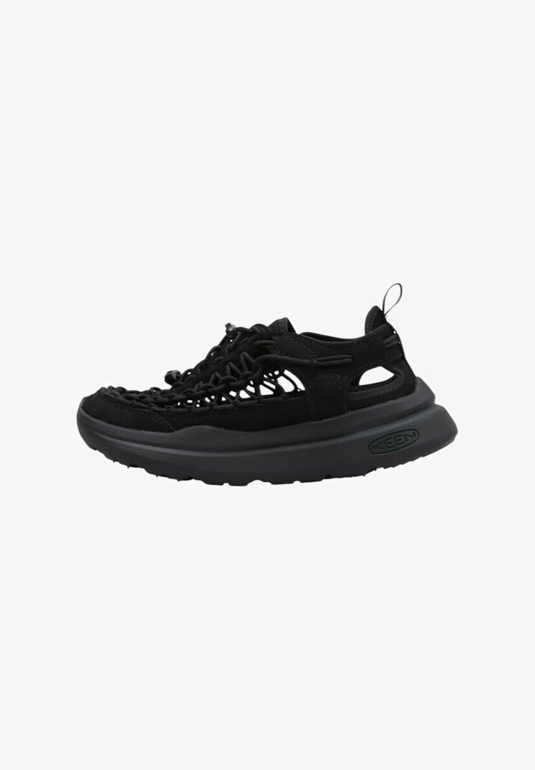 Zapatilla negra con un upper de malla y ante, que presenta una suela de goma gruesa, un detalle tejido en la parte delantera y cortes en los lados.