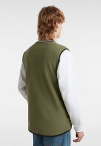 Gilet in pile verde con bordi neri, caratterizzato da un design senza maniche. Indossato sopra una camicia bianca a maniche lunghe e abbinato a jeans in denim blu.