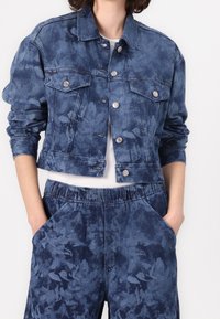 Niebieska kurtka dżinsowa z kwiatowym wzorem, zapinana na guziki, z dwoma kieszeniami na piersiach i dopasowanymi rękawami. Noszona z pasującymi niebieskimi spodenkami dresowymi.