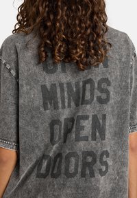 Camiseta gris de gran tamaño hecha de algodón suave, con una textura desgastada y el atrevido texto negro "MENTES ABIERTAS, PUERTAS ABIERTAS" en la parte posterior.