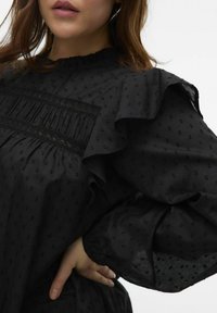 Blusa nera con maniche arricciate, tessuto a pois testurizzati, dettagli plissettati sul petto e colletto alto.