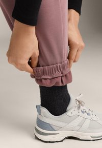 Rózsaszín melegítőnadrág összehúzott szárvégekkel, szürke bordázott zoknival és kék részletekkel díszített fehér futócipővel párosítva. Sima anyagtextúra.