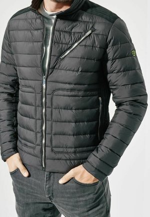 Homme portant une veste matelassée noire avec des poches zippées et les mains dans les poches d'un jean gris foncé sur un fond uni.