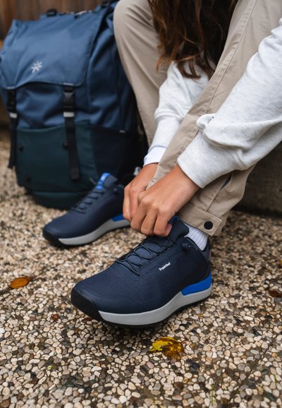 Navy athletic shoes with a textured upper, gray sole, and blue accents; person tying shoelaces while seated on gravel surface.